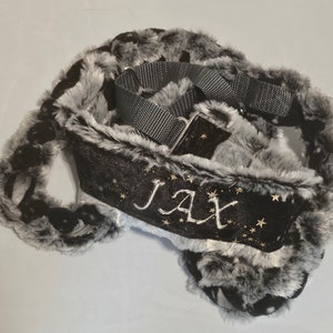 May include: A pet collar featuring a black velvet center panel embroidered with the name JAX in silver thread, surrounded by gold star accents. The collar is bordered by thick, fluffy black and gray faux fur and includes a gray nylon strap.