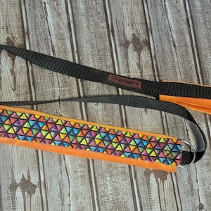 Op de afbeelding: Een zwarte hondenriem met een oranje gevoerde handgreep en een kleurrijk geometrisch patroon op de riem. De riem heeft aan het ene uiteinde twee metalen ringen en de tekst "ALL ROUNDS LLC" is op de handgreep gedrukt.