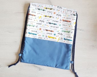 May include: A white drawstring bag with a blue lining. The bag is decorated with a repeating pattern of colorful cartoon race cars and text that says "Cool", "Start", "Finish", "Rally", "Sport", "Go-Go-Go", "Super", "Speed", and "Stop".