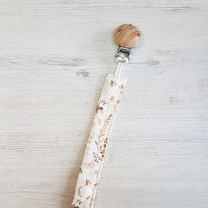 Puede incluir: Un clip para chupete blanco con un patrón floral marrón y un clip de madera. El clip tiene un botón blanco en el extremo.
