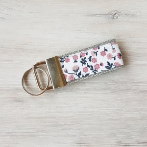 Könnte beinhalten: Ein silberner Schlüsselanhänger mit einem weißen Stoffband mit einem rosafarbenen Blumenmuster.