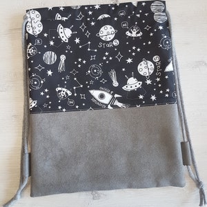 May include: A black drawstring bag with a white space-themed print featuring stars, planets, spaceships, and aliens. The bag has a gray lining and a drawstring closure.