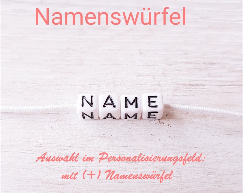 May include: White plastic beads spelling out the word "NAME" on a white string. The text "Auswahl im Personalisierungsfeld: mit (+) Namensw&uuml;rfel" is written in pink below the beads.