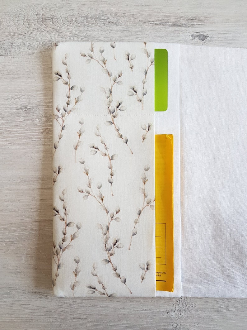 May include: A white fabric envelope with a floral pattern featuring small grey branches and buds. The envelope has a yellow pocket on the right side and a green pocket on the left side.