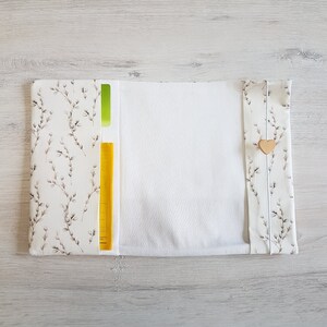 May include: A white fabric book cover with a floral pattern and a yellow pocket. The cover has a white fabric lining and a wooden heart button.