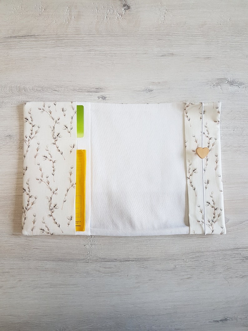 May include: A white fabric book cover with a floral pattern and a yellow pocket. The cover has a white fabric lining and a wooden heart button.