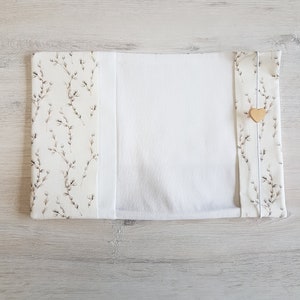 May include: A white book cover with a floral pattern on the front and back. The cover has a white fabric flap that folds over the front and back. A small wooden heart is attached to the flap.