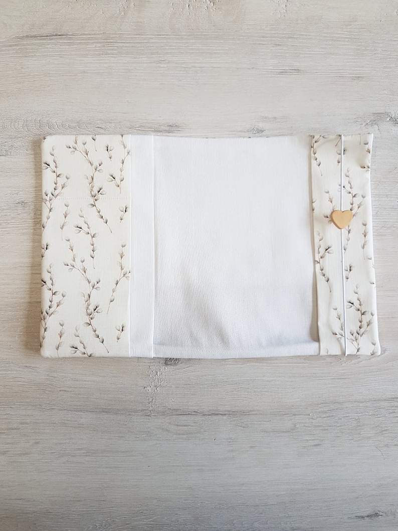 May include: A white book cover with a floral pattern on the front and back. The cover has a white fabric flap that folds over the front and back. A small wooden heart is attached to the flap.