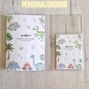 Op de afbeelding: Een U-Heft en Impfpass hoesjesset met een aquarel dinosauruspatroon, palmbomen, eieren en een vulkaan. De tekst luidt U-HEFT en IMPFPASS, gepersonaliseerd met de naam Johann Schneider en de datum 18.07.2025 in een doorzichtige plastic hoes.