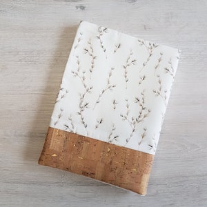 May include: A white fabric book cover with a delicate floral pattern of small white flowers and branches. The cover has a brown cork trim with gold flecks.