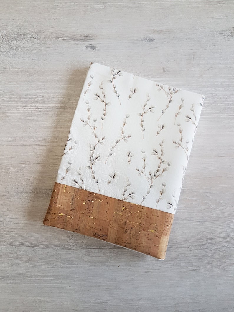 May include: A white fabric book cover with a delicate floral pattern of small white flowers and branches. The cover has a brown cork trim with gold flecks.