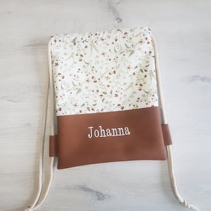 Puede incluir: Mochila con cordón con un estampado floral en la mitad superior y una parte inferior marrón. El nombre "Johanna" está bordado en blanco en la sección marrón. La bolsa tiene correas de cuerda beige.