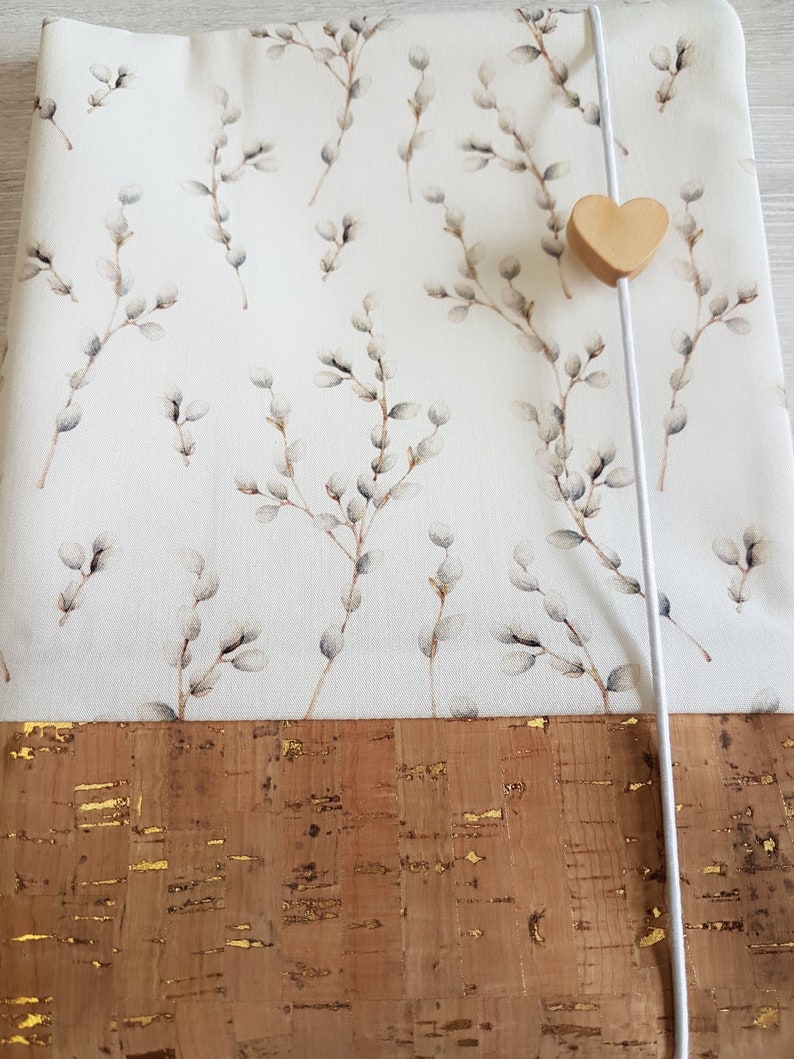 May include: A white fabric notebook cover with a delicate willow branch pattern. The cover has a brown cork bottom with gold flecks and a white ribbon closure with a wooden heart charm.