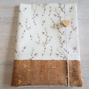 May include: A white fabric laptop sleeve with a floral pattern of willow branches and buds. The sleeve has a brown cork bottom with gold flecks and a white elastic closure with a wooden heart charm.