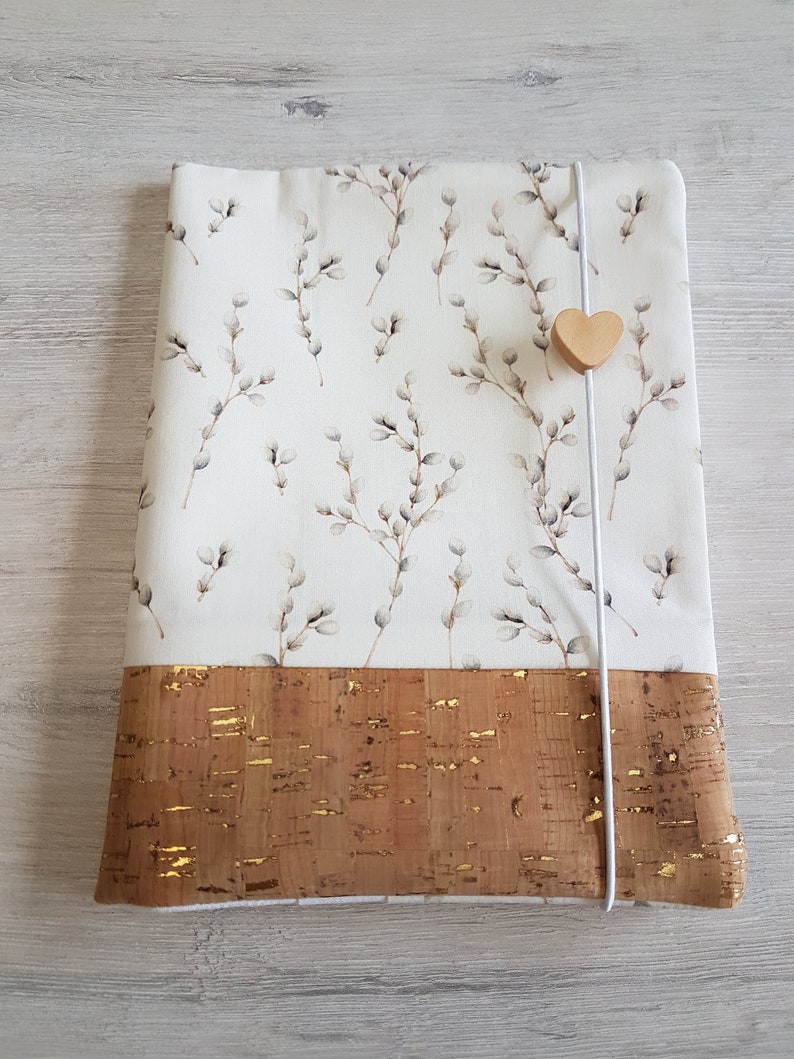 May include: A white fabric laptop sleeve with a floral pattern of willow branches and buds. The sleeve has a brown cork bottom with gold flecks and a white elastic closure with a wooden heart charm.