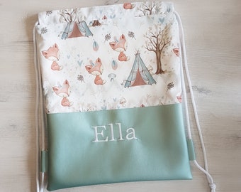Könnte beinhalten: Eine grüne Kordeltasche mit einem weißen Stoffoberteil, das ein Waldmuster mit Füchsen, Bäumen und Zelten zeigt. Die Tasche hat den Namen "Ella" auf der Vorderseite gestickt.