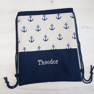 May include: A navy blue drawstring bag with a beige fabric front featuring a repeating pattern of navy blue anchors. The bag has a navy blue pocket on the front with the name "Theodor" embroidered in white.