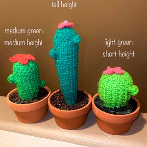 May include: Three crocheted cactus plants in terracotta pots. The cactus on the left is medium green and medium height. The cactus in the middle is dark green and tall. The cactus on the right is light green and short.