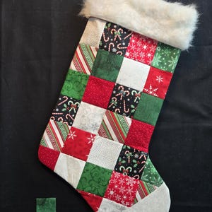 May include: A handmade Christmas stocking with a patchwork design. The stocking features squares of red, green, white, and patterned fabrics with candy canes, snowflakes, and stripes. It has a white faux fur cuff and a gray toe.