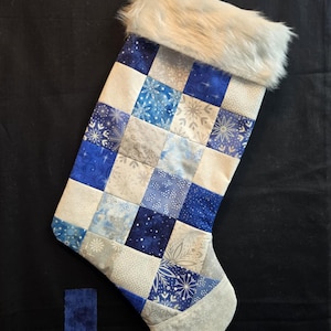 Può includere: Una calza di Natale trapuntata con un design patchwork nei toni del blu, bianco e argento. La calza presenta un bordo in finta pelliccia e motivi a fiocco di neve. Un piccolo quadrato di tessuto blu è accanto alla calza.