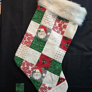 Può includere: Una calza di Natale con un design patchwork con motivi di Babbo Natale, fiocchi di neve e bastoncini di zucchero rossi, verdi e bianchi. La parte superiore della calza è rifinita con ecopelliccia bianca.