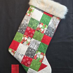 Può includere: Una calza di Natale con un design patchwork con quadrati rossi, verdi, bianchi e grigi. I quadrati mostrano motivi di Babbo Natale, fiocchi di neve e alberi invernali. La parte superiore ha un bordo in ecopelliccia bianca.