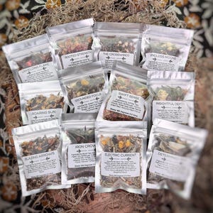May include: An assortment of clear, sealed pouches filled with dried herbs and flowers, each labeled with a unique name. The pouches are arranged on a textured, natural-colored surface, suggesting a collection of herbal teas or blends.