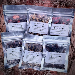 May include: Six small clear plastic bags filled with loose leaf tea blends. Each bag has a white label with a black border and a name for the tea blend. The names are "The Red Death", "Black Cat Yowl", "Beating Heart", "Broken Home", "Quoth the Raven", and "Pendulum Swing".