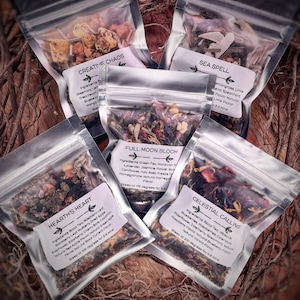 May include: Six small silver pouches containing loose leaf tea blends. Each pouch has a label with the name of the tea blend and a list of ingredients. The tea blends are: Creative Chaos, Sea Spell, Full Moon Bloom, Hearth's Heart, Celestial Calling.
