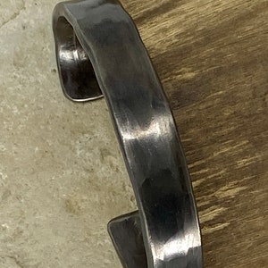 May include: A handcrafted, dark grey metal cuff bracelet. The bracelet has a hammered texture and a slightly curved design. The open ends of the cuff are squared off. The bracelet is shown on a light-coloured surface.