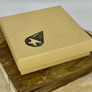 Puede incluir: Una caja de cart&oacute;n marr&oacute;n con un logotipo de gota de agua negra. La caja est&aacute; sobre una superficie de madera.