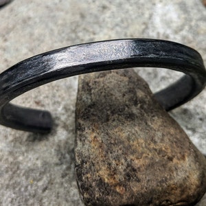 Puede incluir: Un brazalete de metal negro con un acabado liso y pulido. El brazalete est&aacute; hecho de un metal grueso y resistente y tiene un dise&ntilde;o simple y minimalista.