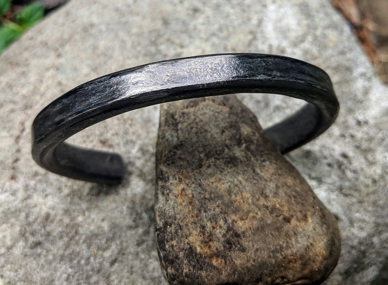 Puede incluir: Un brazalete de metal negro con un acabado liso y pulido. El brazalete est&aacute; hecho de un metal grueso y resistente y tiene un dise&ntilde;o simple y minimalista.