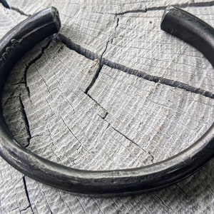 Puede incluir: Un brazalete de metal negro grueso con una superficie redonda y lisa. El brazalete est&aacute; sobre una superficie de madera clara.