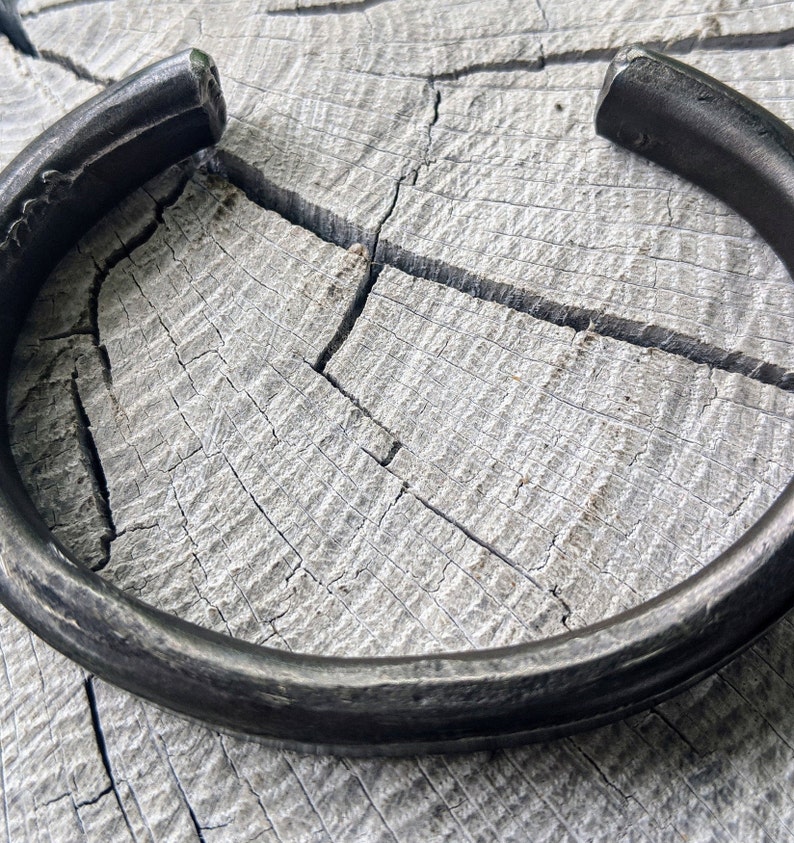 Puede incluir: Un brazalete de metal negro grueso con una superficie redonda y lisa. El brazalete est&aacute; sobre una superficie de madera clara.