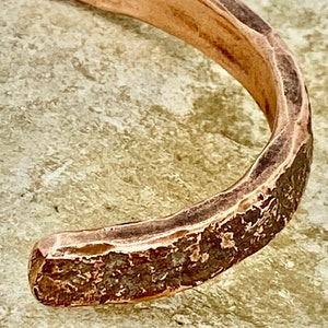 K&ouml;nnte beinhalten: Nahaufnahme eines Kupferarmbands mit geh&auml;mmerter Textur. Das Armband ist schlicht und minimalistisch gestaltet mit einer abgerundeten Form.