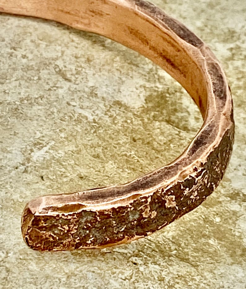 K&ouml;nnte beinhalten: Nahaufnahme eines Kupferarmbands mit geh&auml;mmerter Textur. Das Armband ist schlicht und minimalistisch gestaltet mit einer abgerundeten Form.