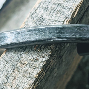 Puede incluir: Un brazalete de metal plateado con acabado martillado. El brazalete es curvo y tiene una superficie lisa y pulida.