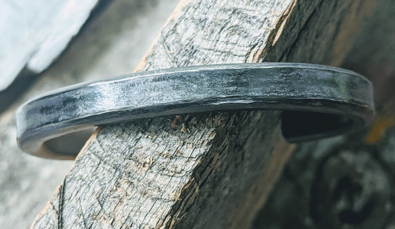 Puede incluir: Un brazalete de metal plateado con acabado martillado. El brazalete es curvo y tiene una superficie lisa y pulida.