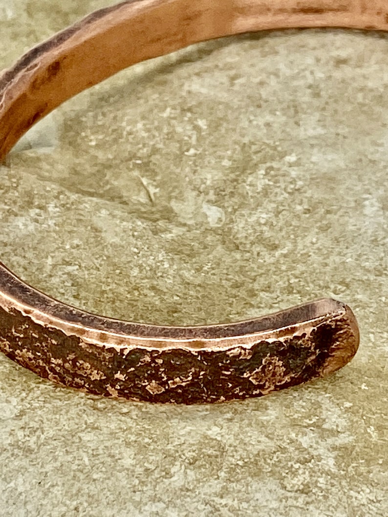 K&ouml;nnte beinhalten: Nahaufnahme eines Kupferarmbands mit geh&auml;mmerter Textur. Das Armband ist offen und hat eine leicht abgerundete Form.