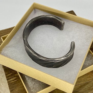 Puede incluir: Un brazalete de metal plateado con un dise&ntilde;o texturizado y trenzado. El brazalete se muestra en una caja marr&oacute;n con papel de seda blanco.