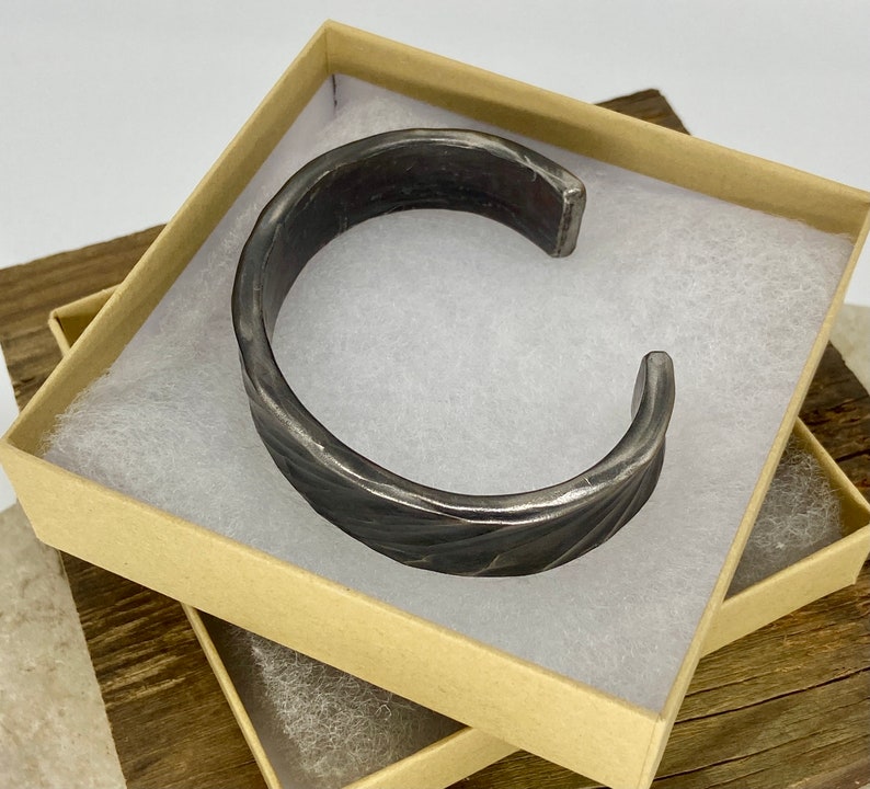 Puede incluir: Un brazalete de metal plateado con un dise&ntilde;o texturizado y trenzado. El brazalete se muestra en una caja marr&oacute;n con papel de seda blanco.