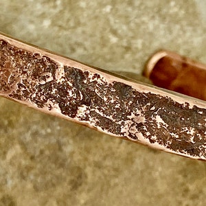 May include: A close-up of a copper bracelet with a hammered texture. The bracelet is made of a single piece of metal and has a rustic, aged look.