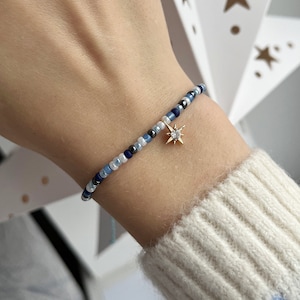 Puede incluir: Un brazalete de cuentas azules y blancas con un dije de estrella dorada. La estrella tiene una pequeña piedra transparente en el centro.