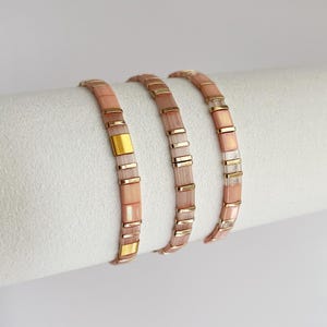 Puede incluir: Tres pulseras de cuentas rosas y doradas. Las pulseras están hechas de pequeñas cuentas rectangulares y detalles metálicos dorados. Se exhiben sobre una superficie blanca. Cada pulsera mide aproximadamente 18 cm.
