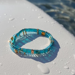May include: A stretch bracelet composed of translucent turquoise blue rectangular glass beads interspersed with small metallic gold-toned cylindrical spacers. The beads are arranged in a single continuous loop, creating a vibrant, reflective accessory.