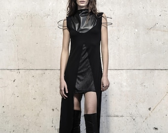 Puede incluir: Un vestido negro sin mangas con cuello alto y panel liso, una abertura frontal que revela una capa inferior texturizada y finos detalles de alambre circular en los hombros. La prenda se combina con botas altas de cuero negro.