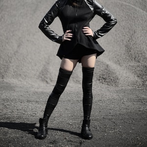 Puede incluir: Una mujer con una chaqueta de cuero negra con cierre de cremallera, medias negras hasta los muslos y botas negras. Está de pie frente a un fondo de grava gris.