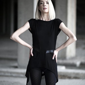 May include: A black tunic-style top with short sleeves and a high-low hemline. The top features leather-like accents and is paired with black leggings. The model has blonde hair and is standing in a concrete environment.