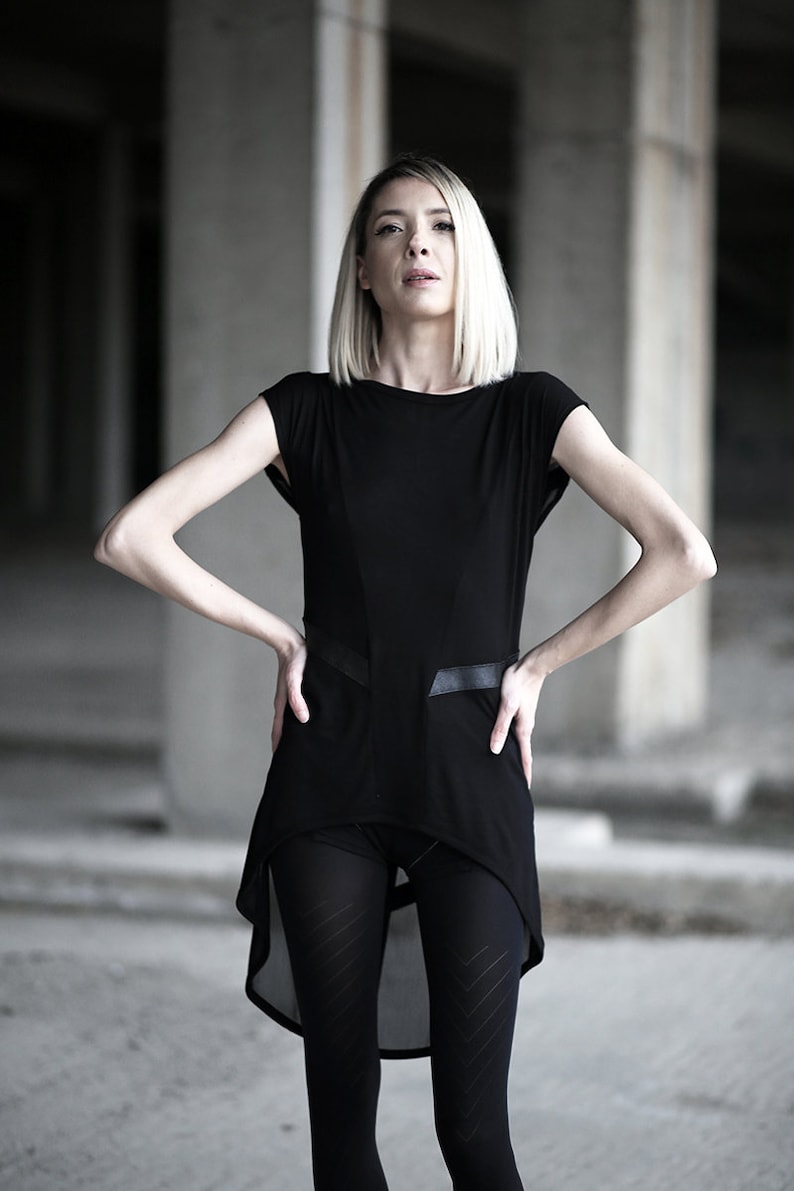 May include: A black tunic-style top with short sleeves and a high-low hemline. The top features leather-like accents and is paired with black leggings. The model has blonde hair and is standing in a concrete environment.